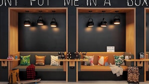 A modern room with a black wall, wooden benches, and pendant lights. There are pillows, a skateboard, and a suitcase on the benches. - Moxy Milton Keynes (Milton Keynes)