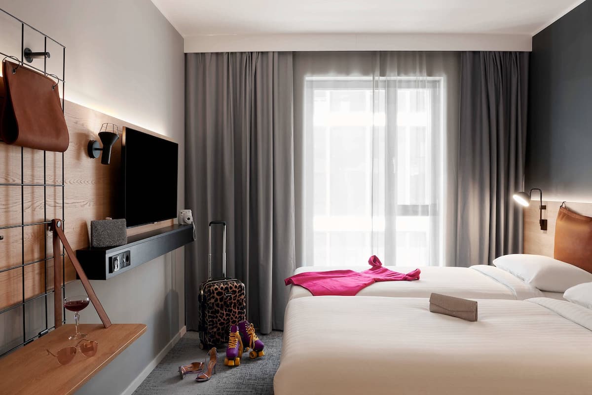 Habitación de hotel con una cama grande, un televisor montado en la pared, una maleta y una mesita con una copa de vino y unas gafas de sol.