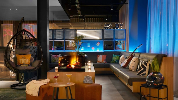 A modern lounge area with a hanging chair, a sofa, a small table with a drink, and a decorative wall with lights. - Moxy Milton Keynes (Milton Keynes)