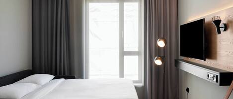 Habitación de hotel con cama, televisor montado en la pared, mesita de noche y ventana con cortinas.