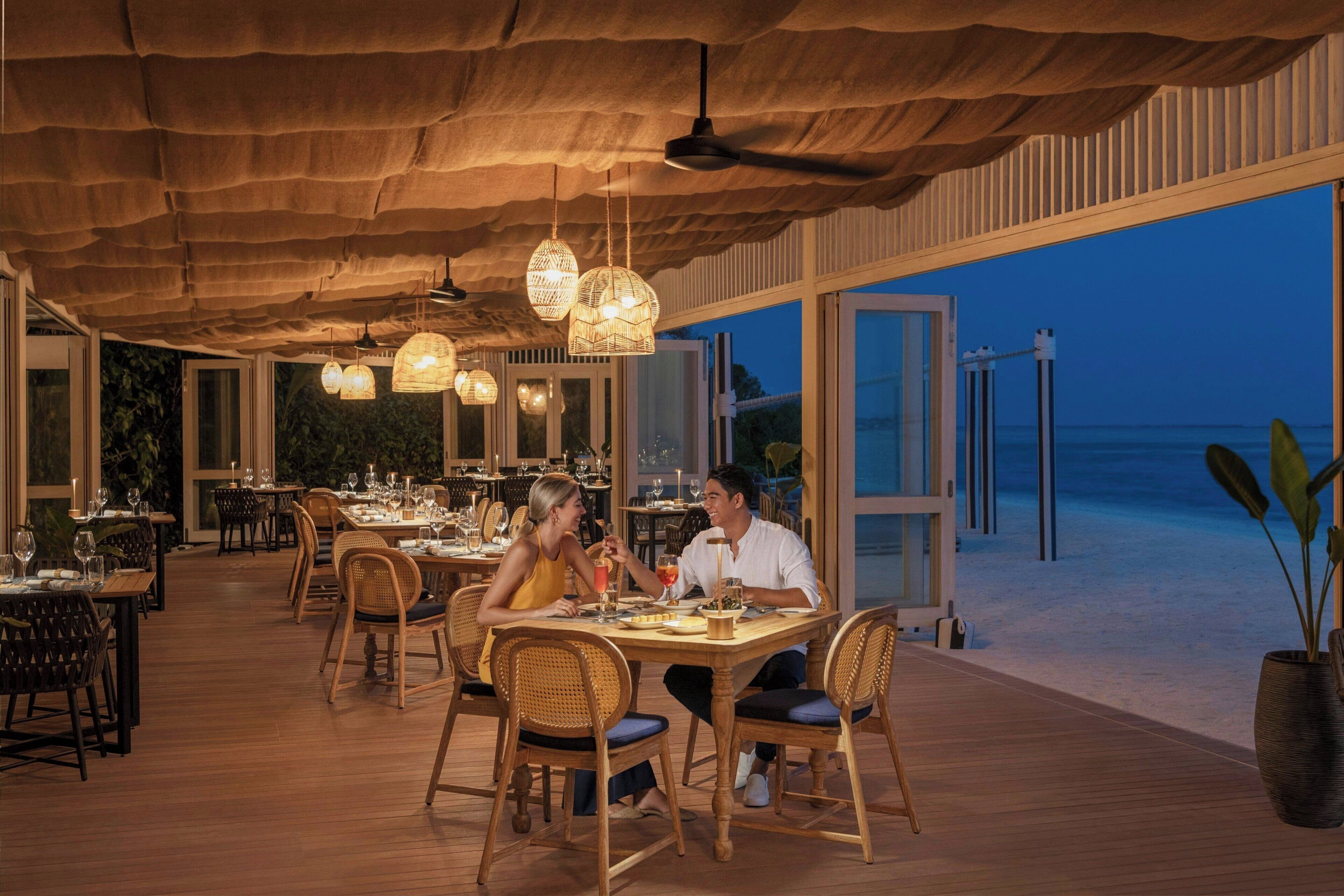 An outdoor dining area with tables and chairs, large windows, and a view of the ocean.