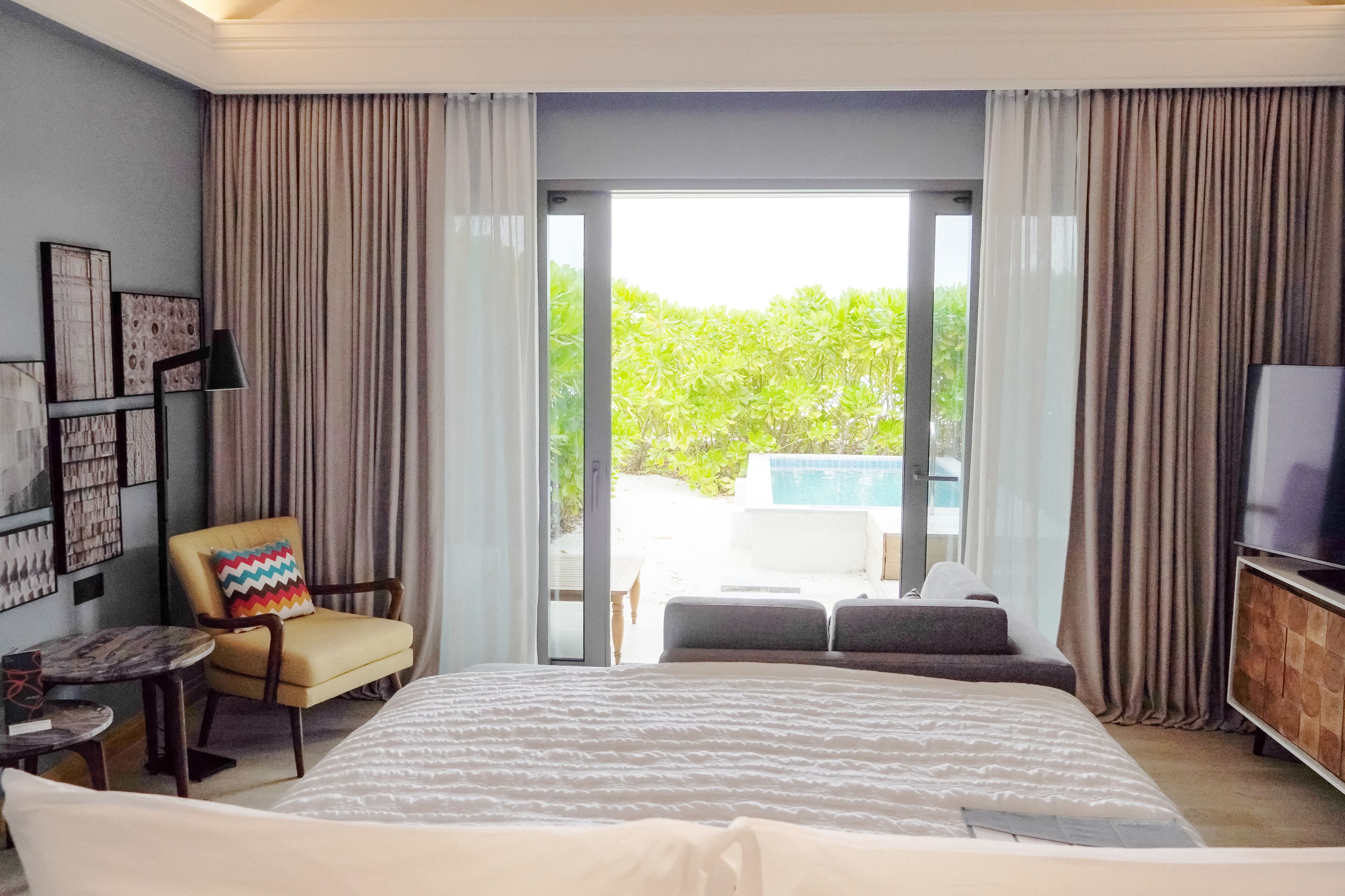 Une chambre d’hôtel moderne dotée d’un grand lit, d’un coin salon avec un fauteuil et un canapé, d’une télévision et d’un balcon offrant une vue sur la piscine.