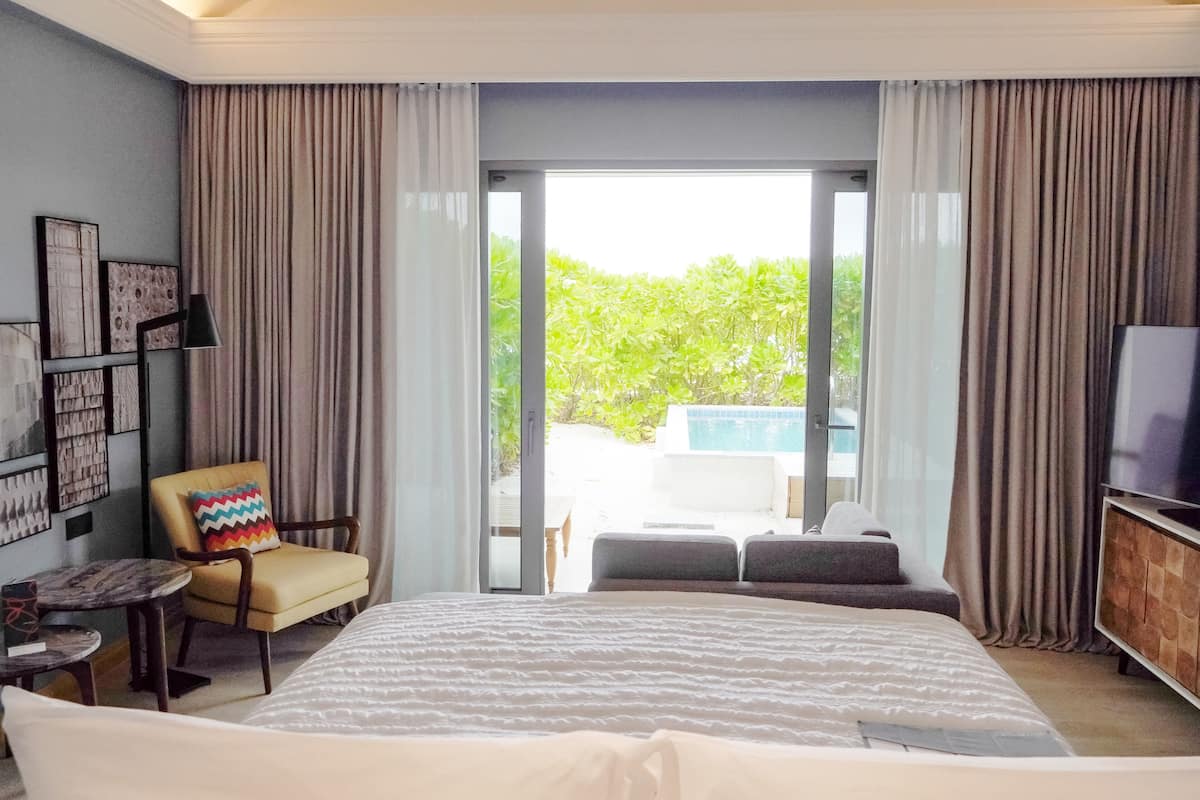 Una habitación de hotel moderna con una cama grande, una zona de estar con una silla y un sofá, televisión y un balcón con vistas a la piscina.