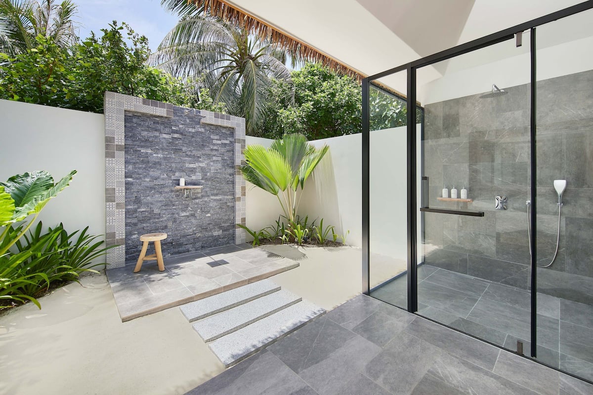 Un área de ducha exterior moderna con puerta de cristal, pared de piedra y un taburete de madera pequeño.