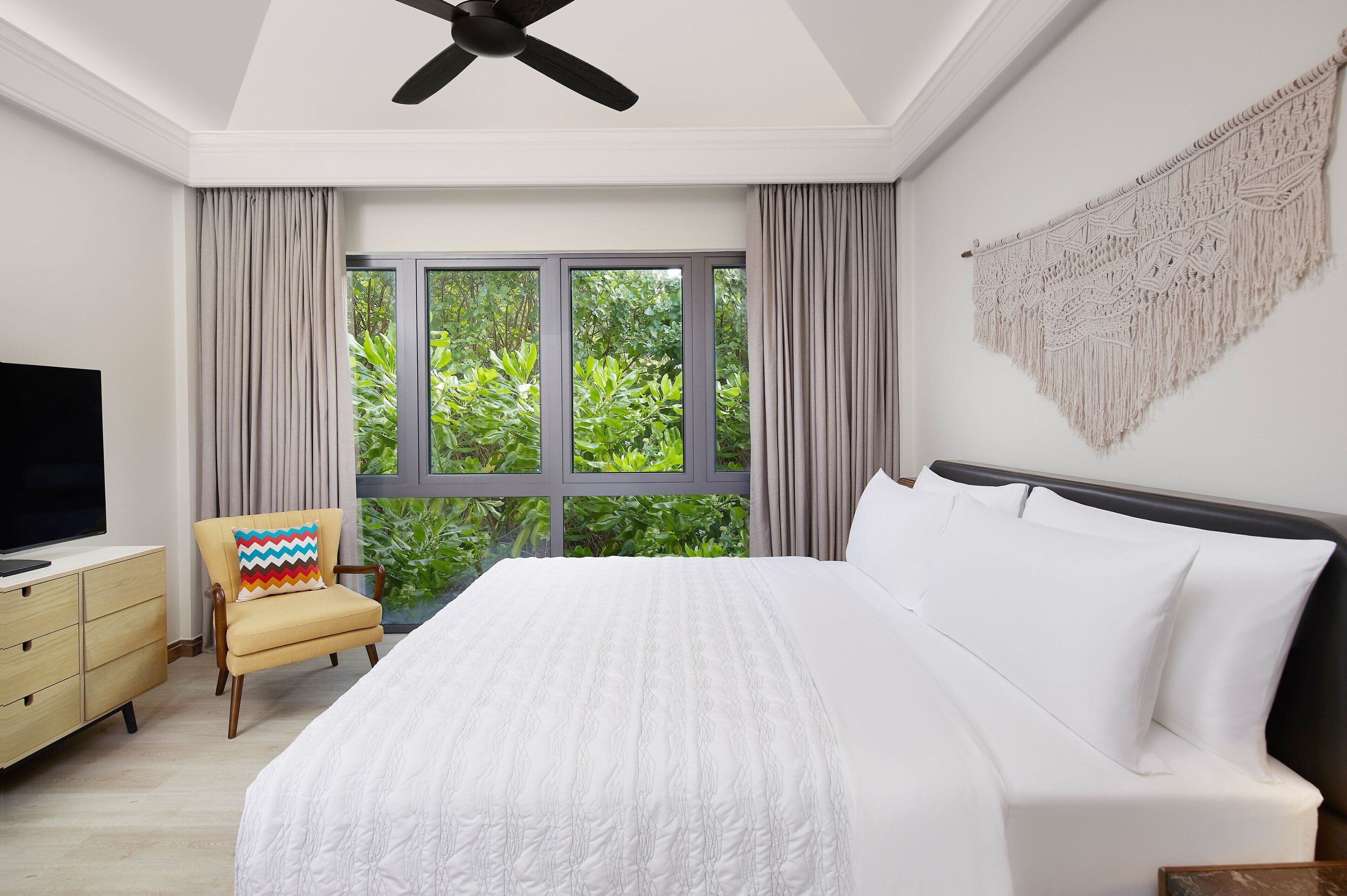 Une chambre avec un grand lit, un téléviseur posé sur un meuble en bois, une chaise avec un coussin coloré et une vue sur la verdure par la fenêtre.