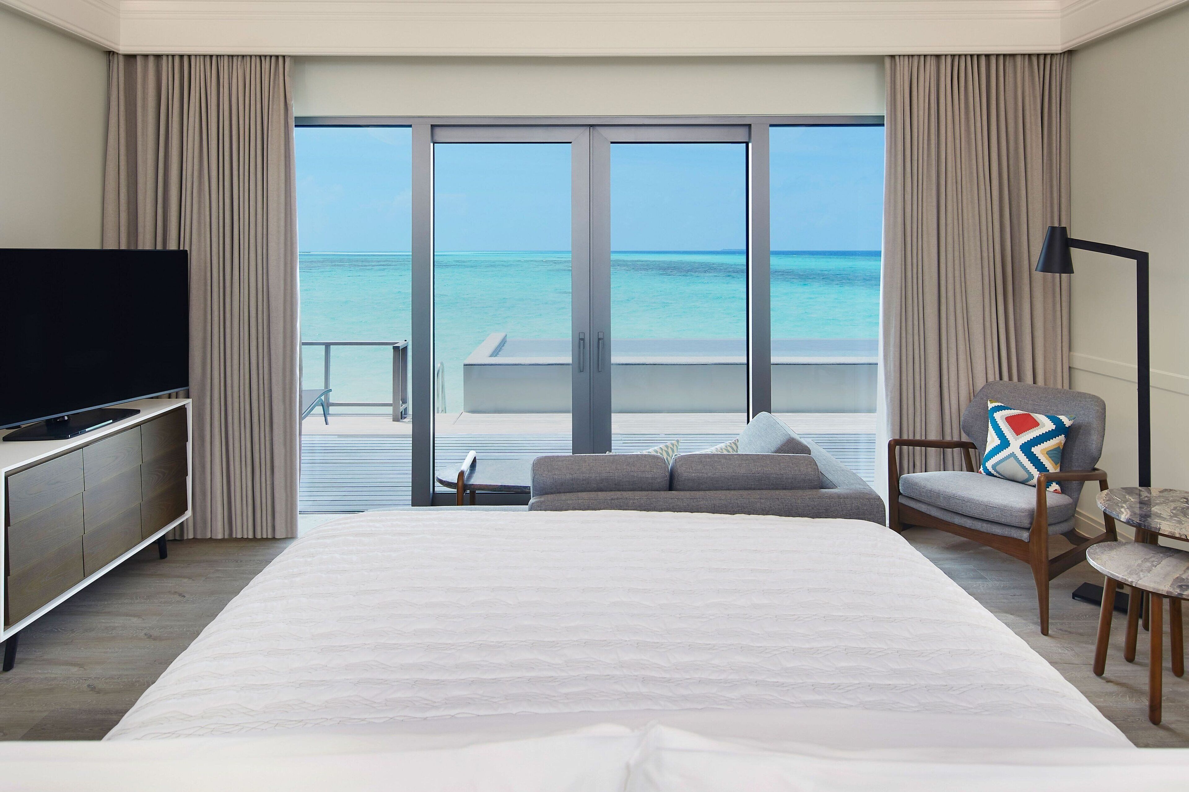 A hotel room with a large bed, a TV, a chair, and a view of the ocean through large windows.