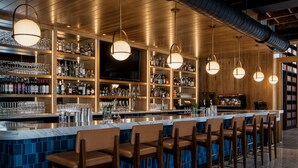 Uma área de bar com um balcão longo, várias prateleiras com garrafas, copos e uma tela de televisão.