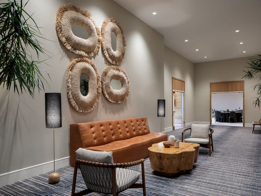 Een hal met een bruine leren bank, een ronde houten salontafel en een rieten stoel. Er zijn vier grote geweven wanddecoraties, een hoge vloerlamp en een potplant.
