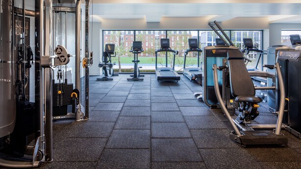 A gym with cardio machines, weight training equipment, and a view of a building through large windows. - The Ellison, Oklahoma City, a Tribute Portfolio Hotel (Oklahoma City)