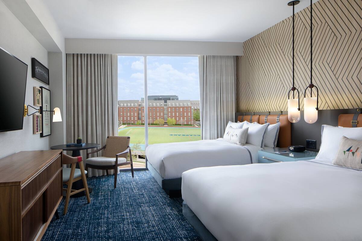 A hotel room with two beds, a desk, a chair, and a large window with a view of a building.