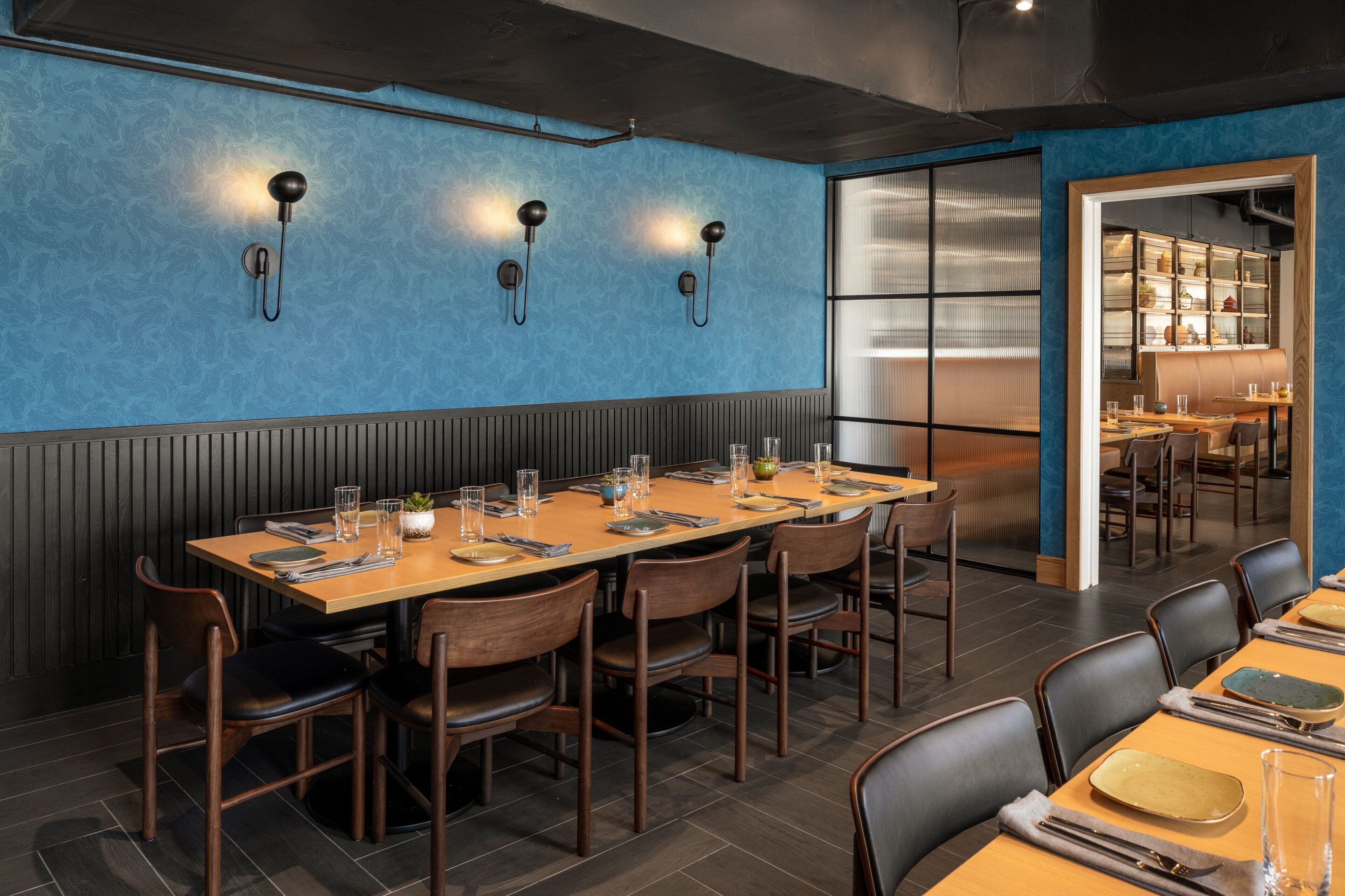 Un restaurant doté d’une longue table en bois dressée pour le repas, d’un mur bleu à la texture particulière et d’un espace cuisine visible à travers une cloison en verre.