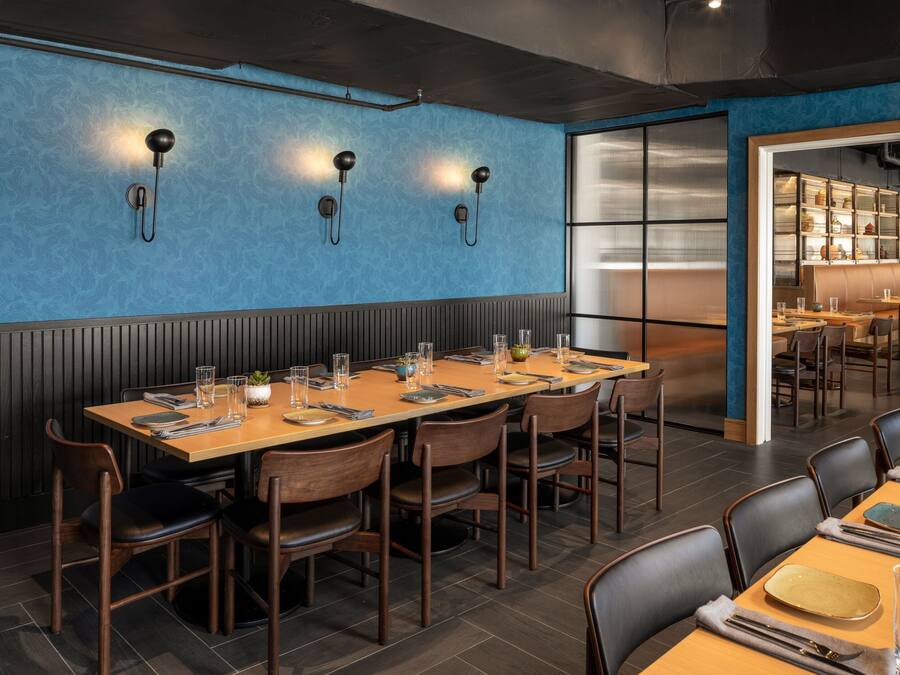 Un restaurant doté d’une longue table en bois dressée pour le repas, d’un mur bleu à la texture particulière et d’un espace cuisine visible à travers une cloison en verre.