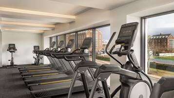 A row of treadmills in a gym with large windows and a view of buildings outside.