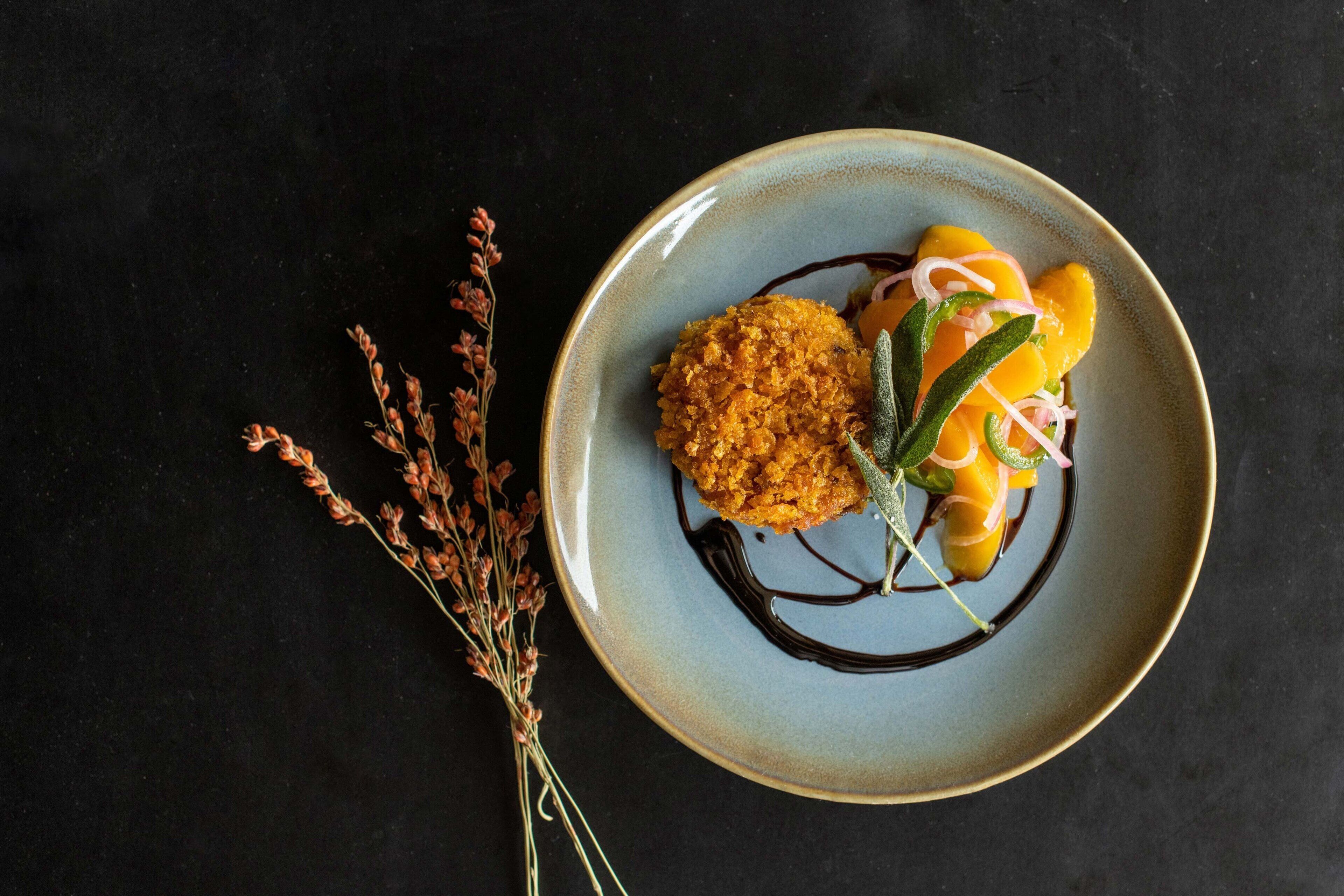 Une assiette contenant un plat, peut-être un dessert, agrémenté d’herbes et nappé d’une sauce, accompagnée d’une branche de fleurs séchées sur un fond sombre.