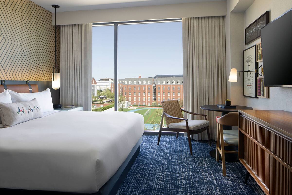 A hotel room with a large bed, a desk with a chair, a TV, and a view of a building through large windows.