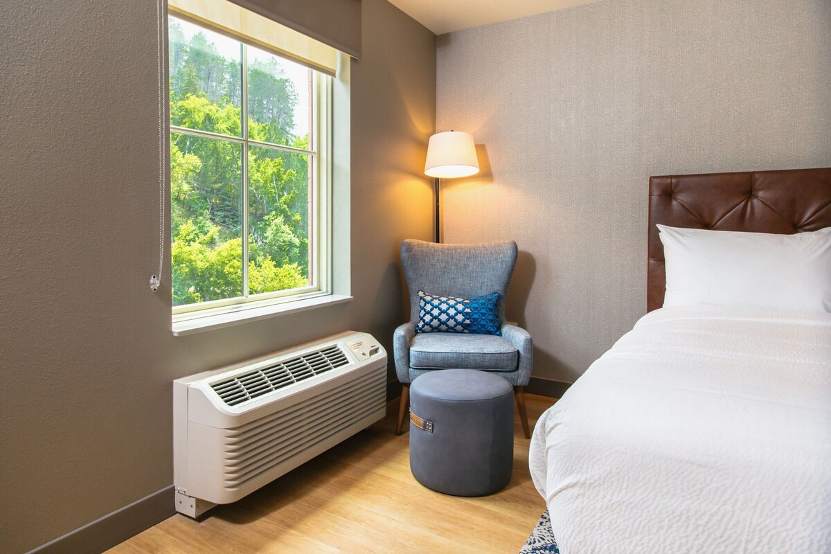 Une chambre d’hôtel avec un lit, une chaise avec un coussin, un lampadaire et une fenêtre donnant sur des arbres.