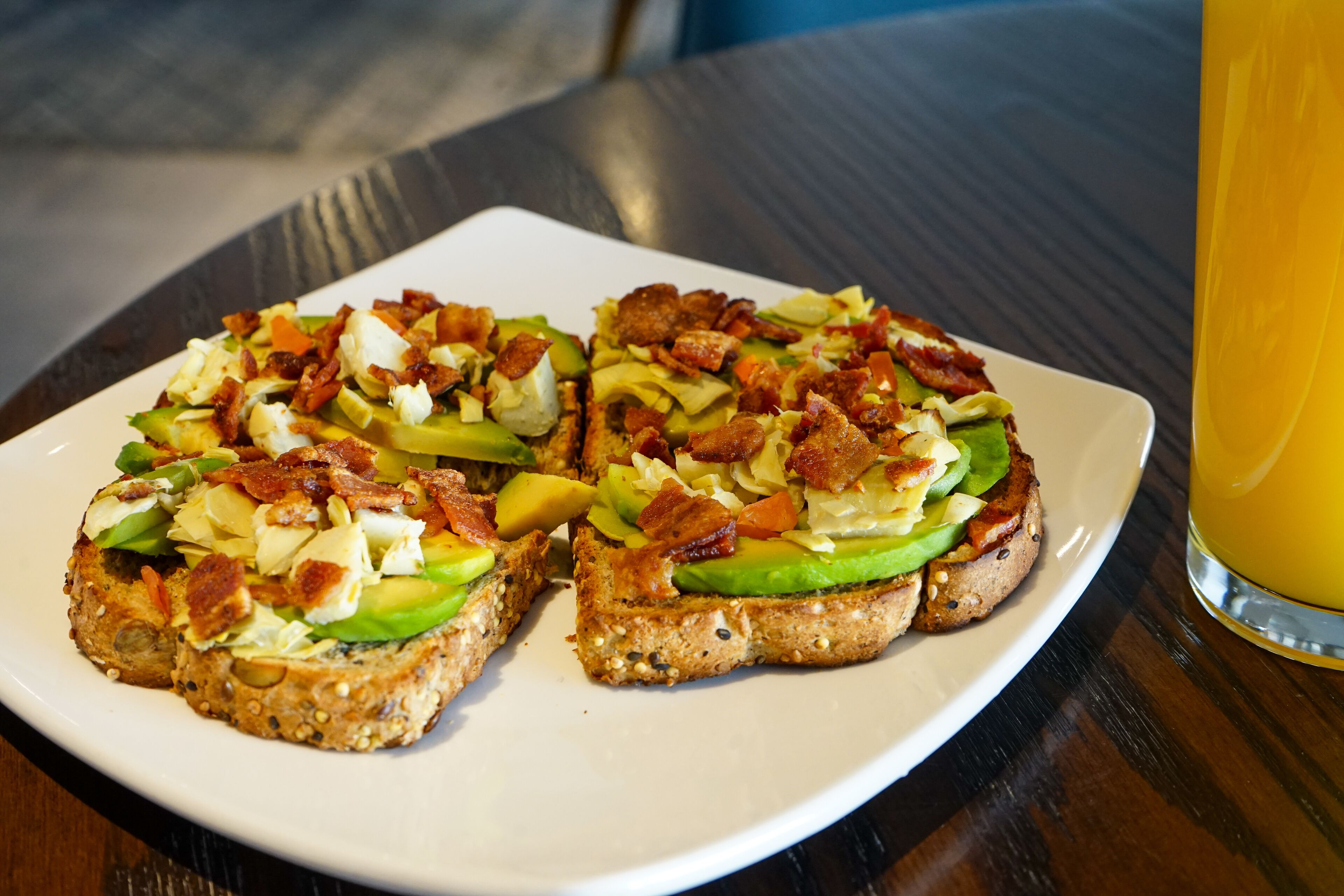 A plate with two slices of toast topped with avocado, bacon, and cheese, and a glass of orange juice on a wooden table.