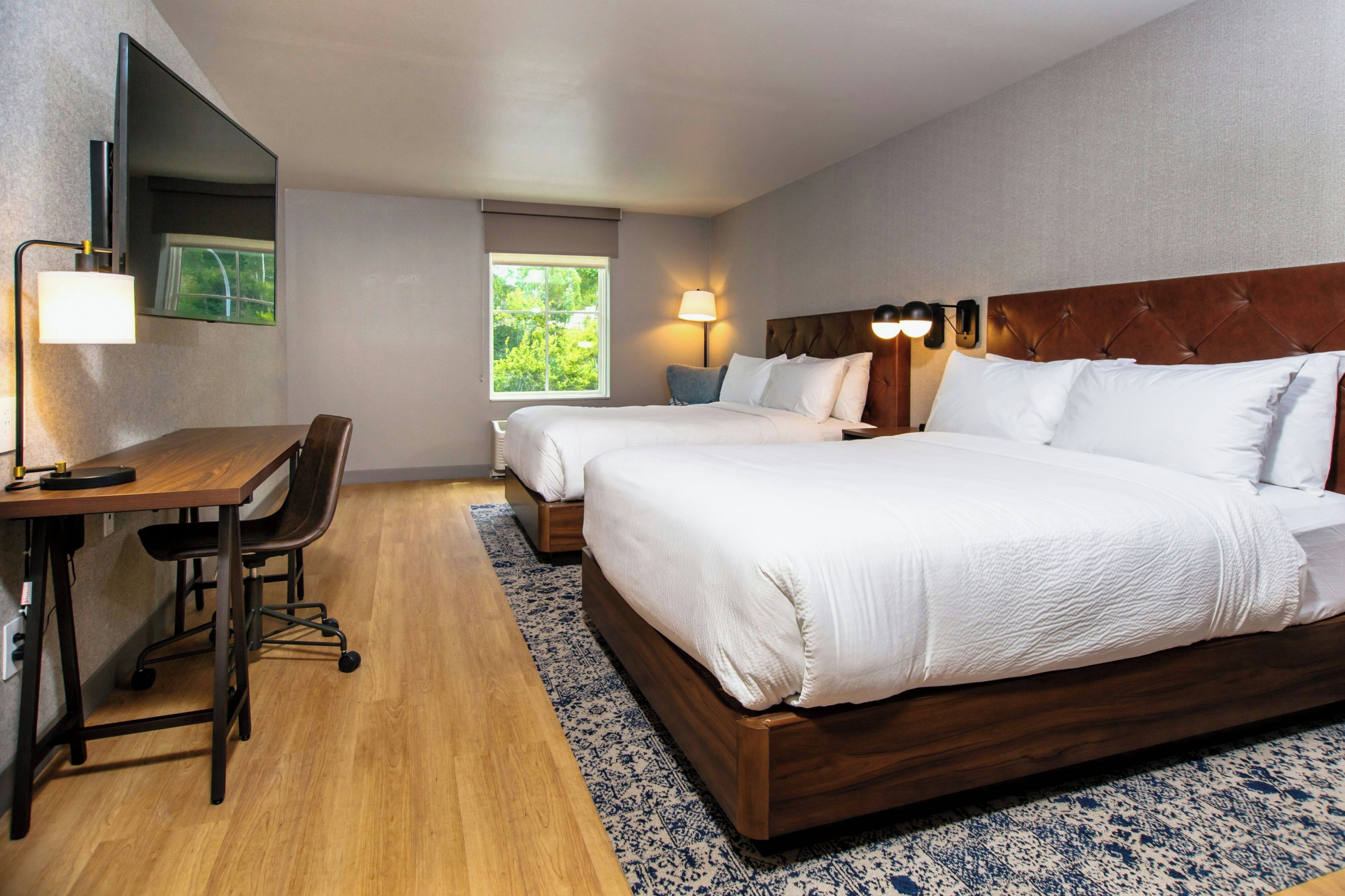 Une chambre d’hôtel avec deux lits, un bureau, une chaise, une grande fenêtre et un téléviseur à écran plat.