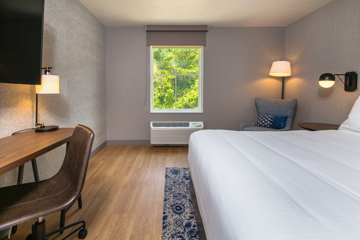 Une chambre d’hôtel équipée d’un lit, d’un bureau, d’une chaise, d’une fenêtre donnant sur des arbres et d’un climatiseur fixé au mur.