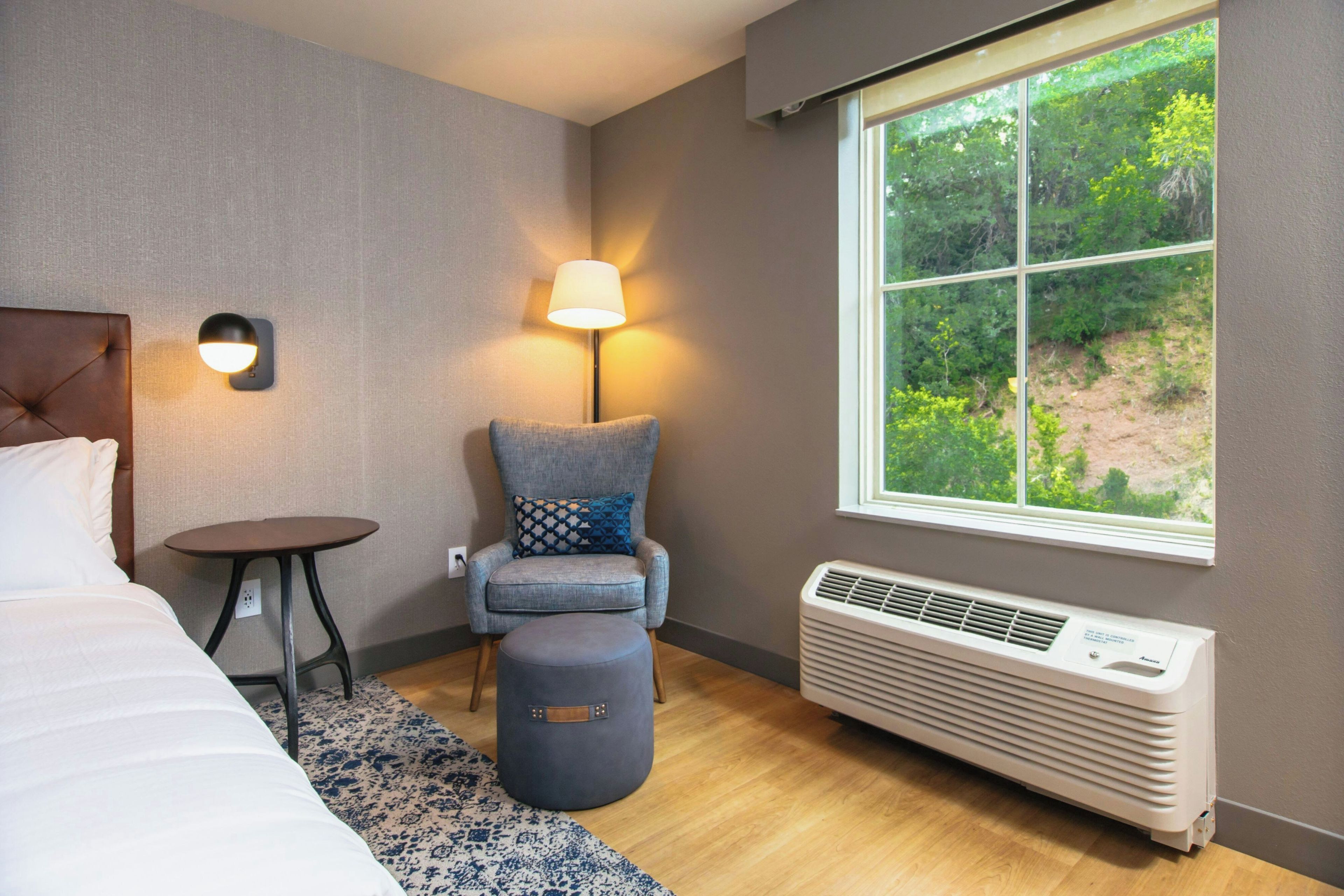 Ein Hotelzimmer mit einem Bett, einem Sessel mit Kissen, einem kleinen Tisch, einer Stehlampe und einem Fenster mit Blick ins Grüne.