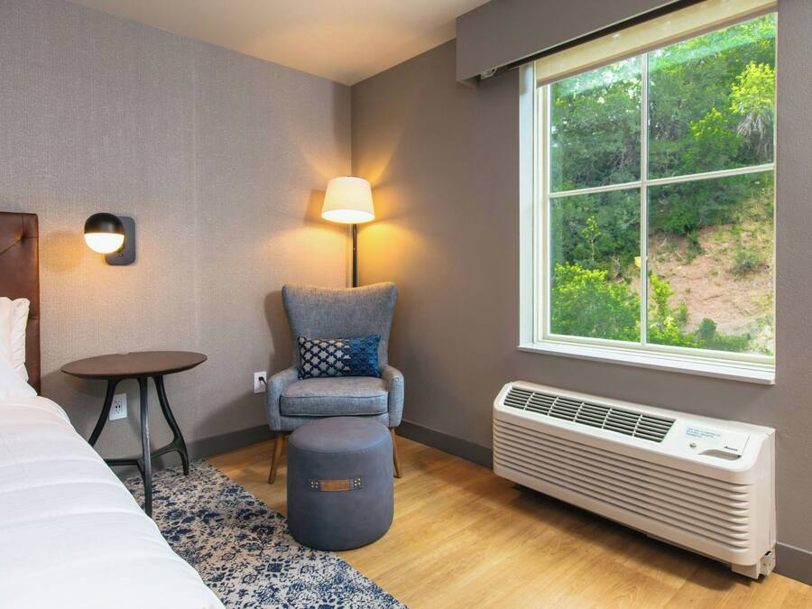 Ein Hotelzimmer mit einem Bett, einem Sessel mit Kissen, einem kleinen Tisch, einer Stehlampe und einem Fenster mit Blick ins Grüne.