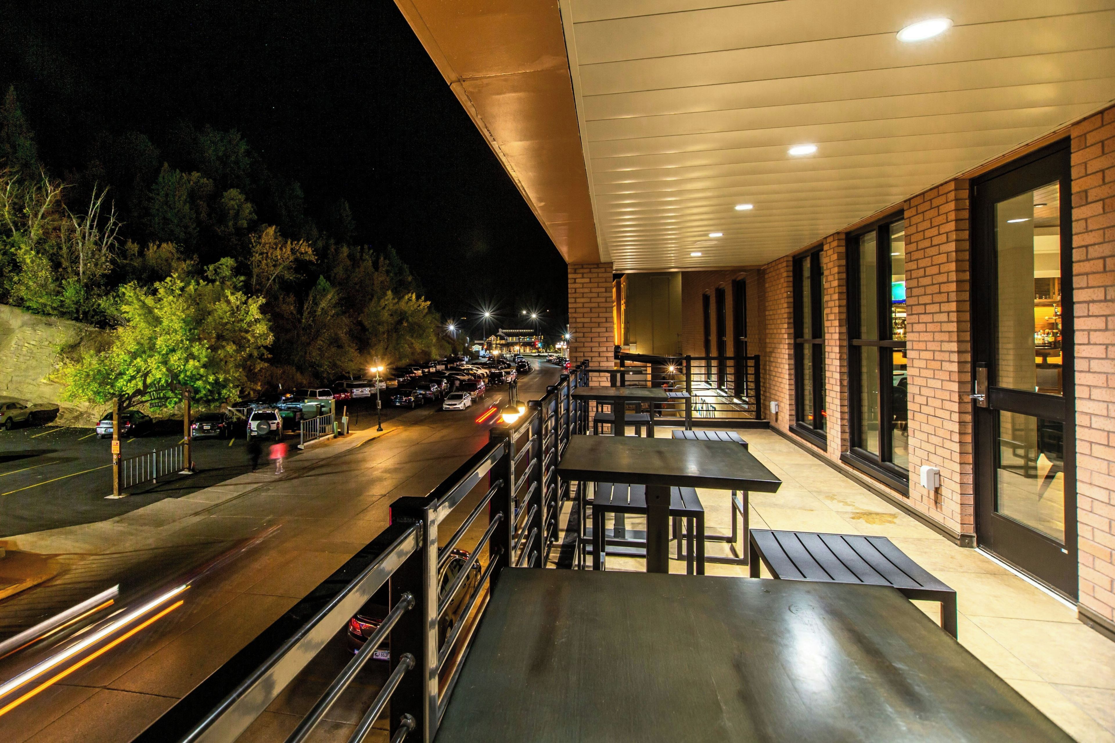 An outdoor seating area with tables and benches, overlooking a street with parked cars and a building with large windows.