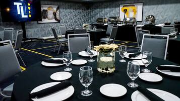 Un salón de conferencias con mesas dispuestas para una reunión, equipado con sillas, platos, vasos y un jarrón decorativo.
