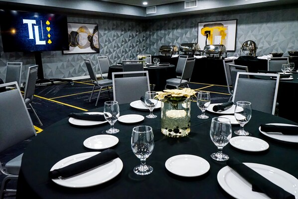 Un salón de conferencias con mesas dispuestas para una reunión, equipado con sillas, platos, vasos y un jarrón decorativo.