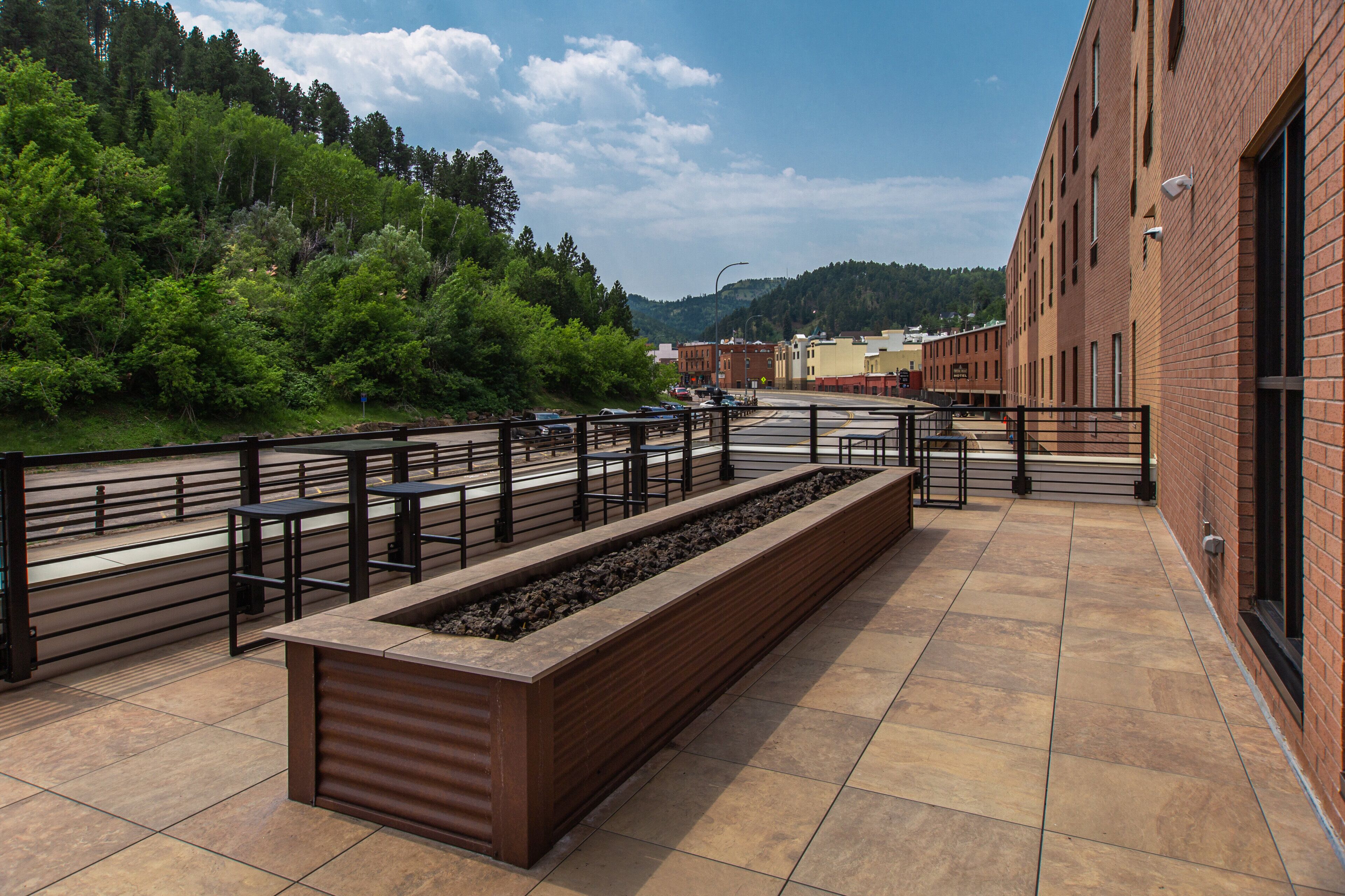 Una terraza exterior con un largo banco, baranda de metal y vistas a una ladera boscosa.