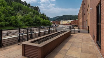 Una terraza exterior con un largo banco, baranda de metal y vistas a una ladera boscosa.