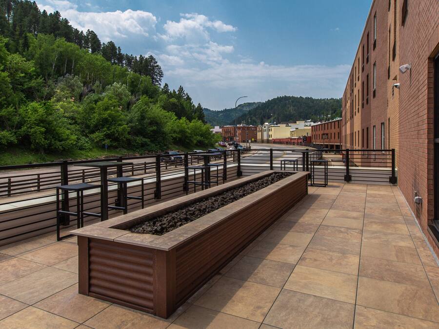Una terraza exterior con un largo banco, baranda de metal y vistas a una ladera boscosa.