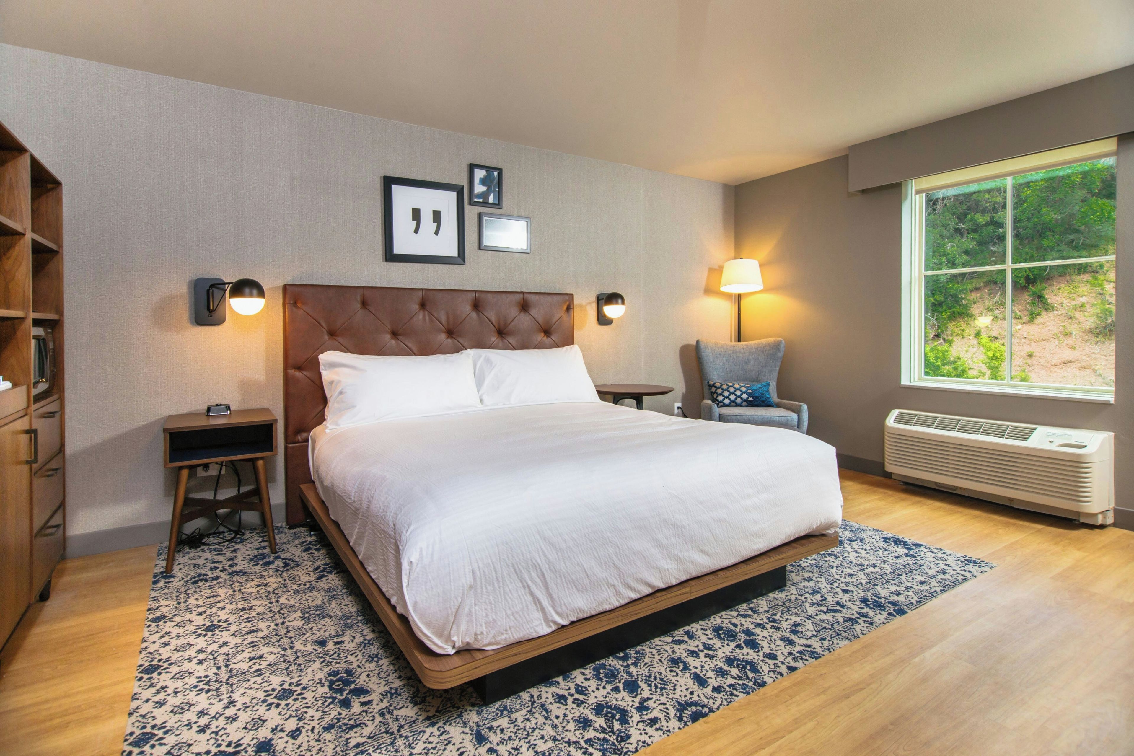 A hotel room with a large bed, a bedside table, a chair, a window with a view, and a wall-mounted lamp.