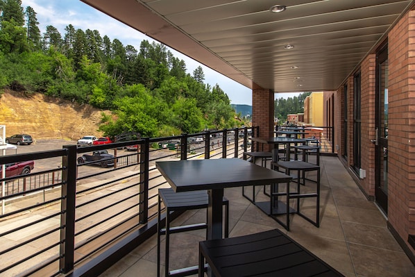 A covered outdoor seating area with tables and chairs, overlooking a road and forested hillside.