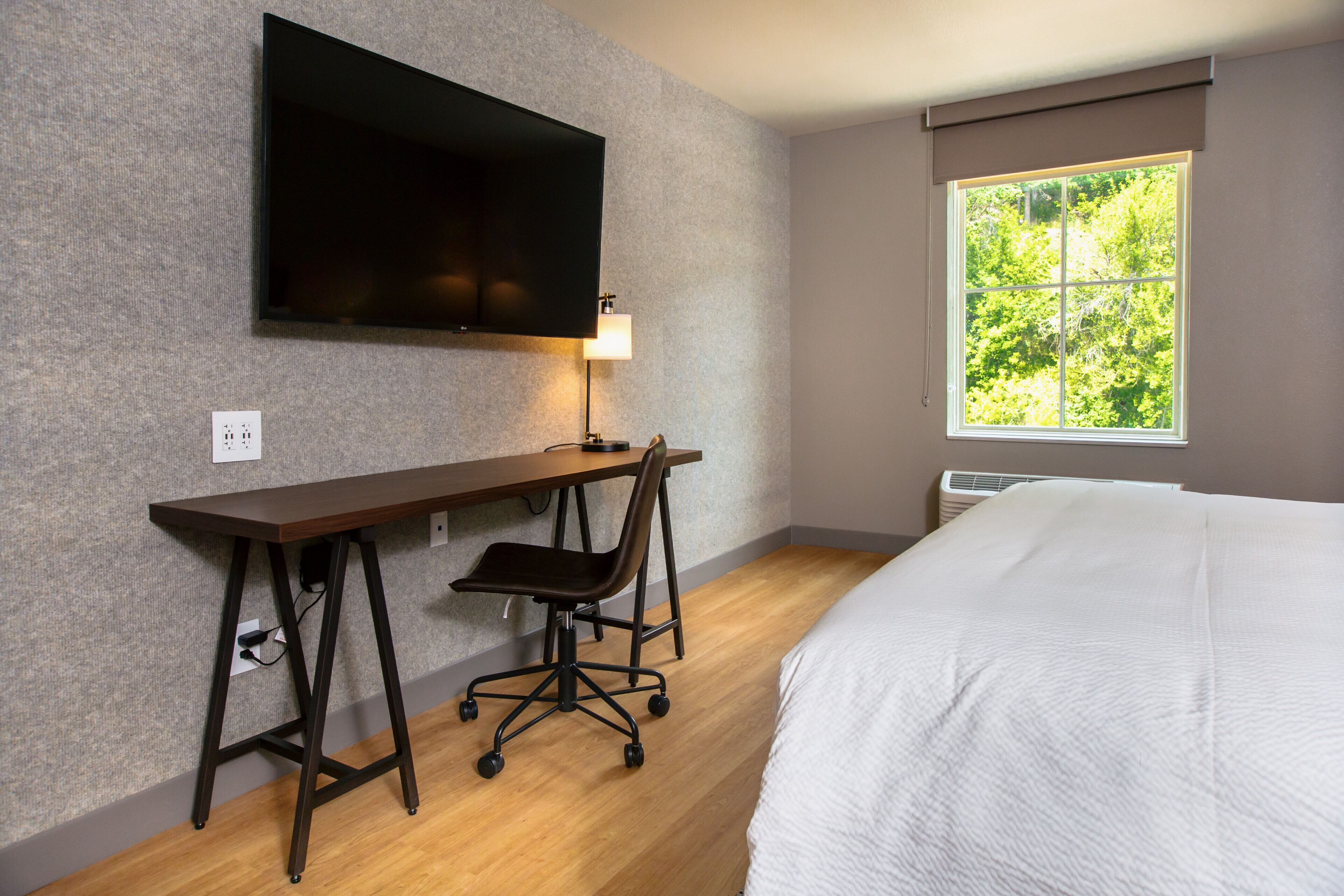 Quarto de hotel com cama, escrivaninha com cadeira, televisão na parede e janela com vista para árvores.