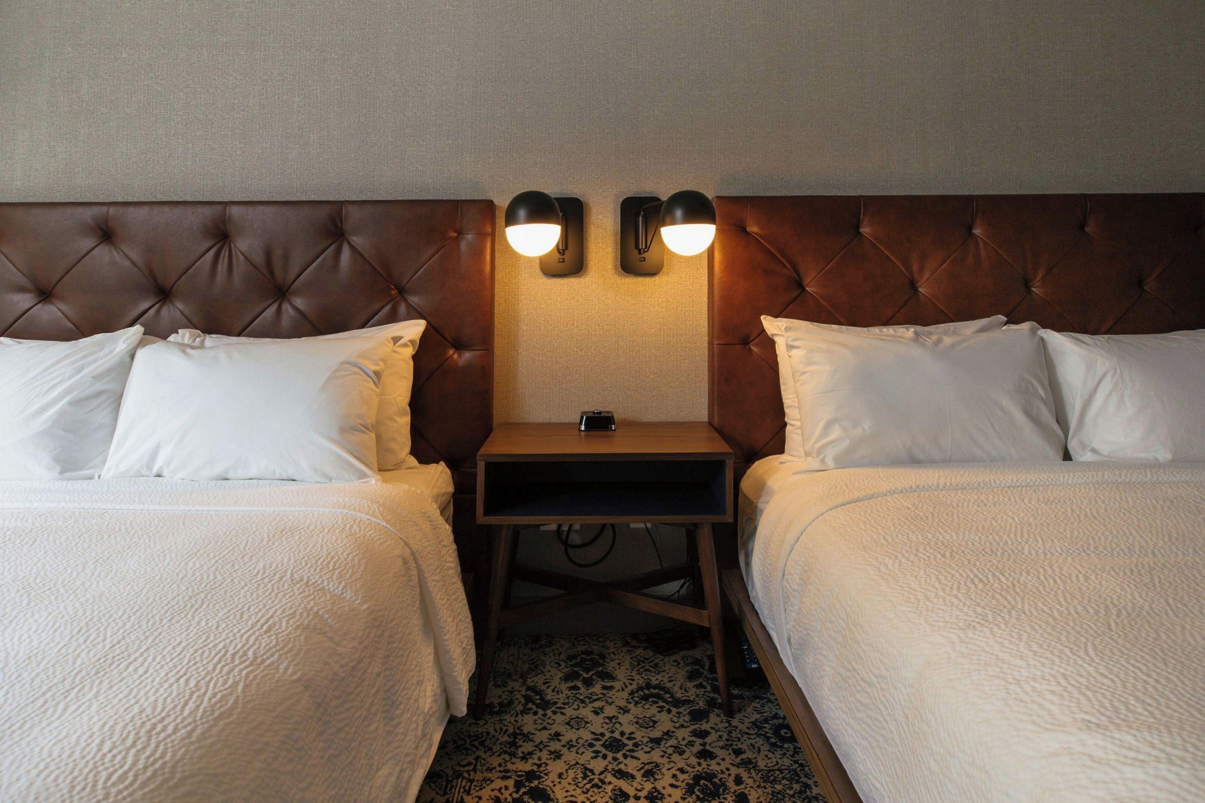 A hotel room with two beds, a nightstand, and two wall-mounted lamps.