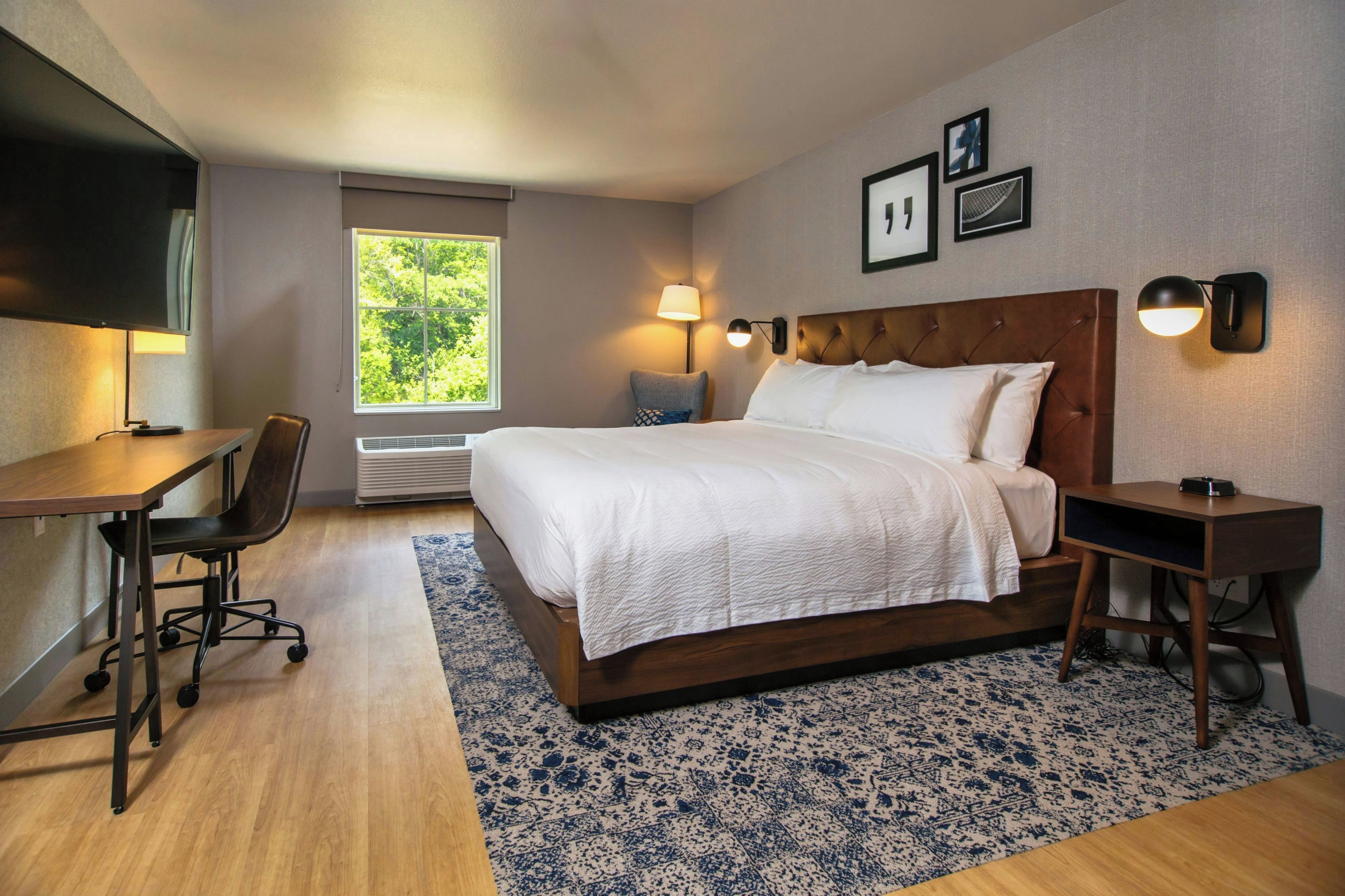 A hotel room with a bed, desk, chair, and a window with a view of greenery.