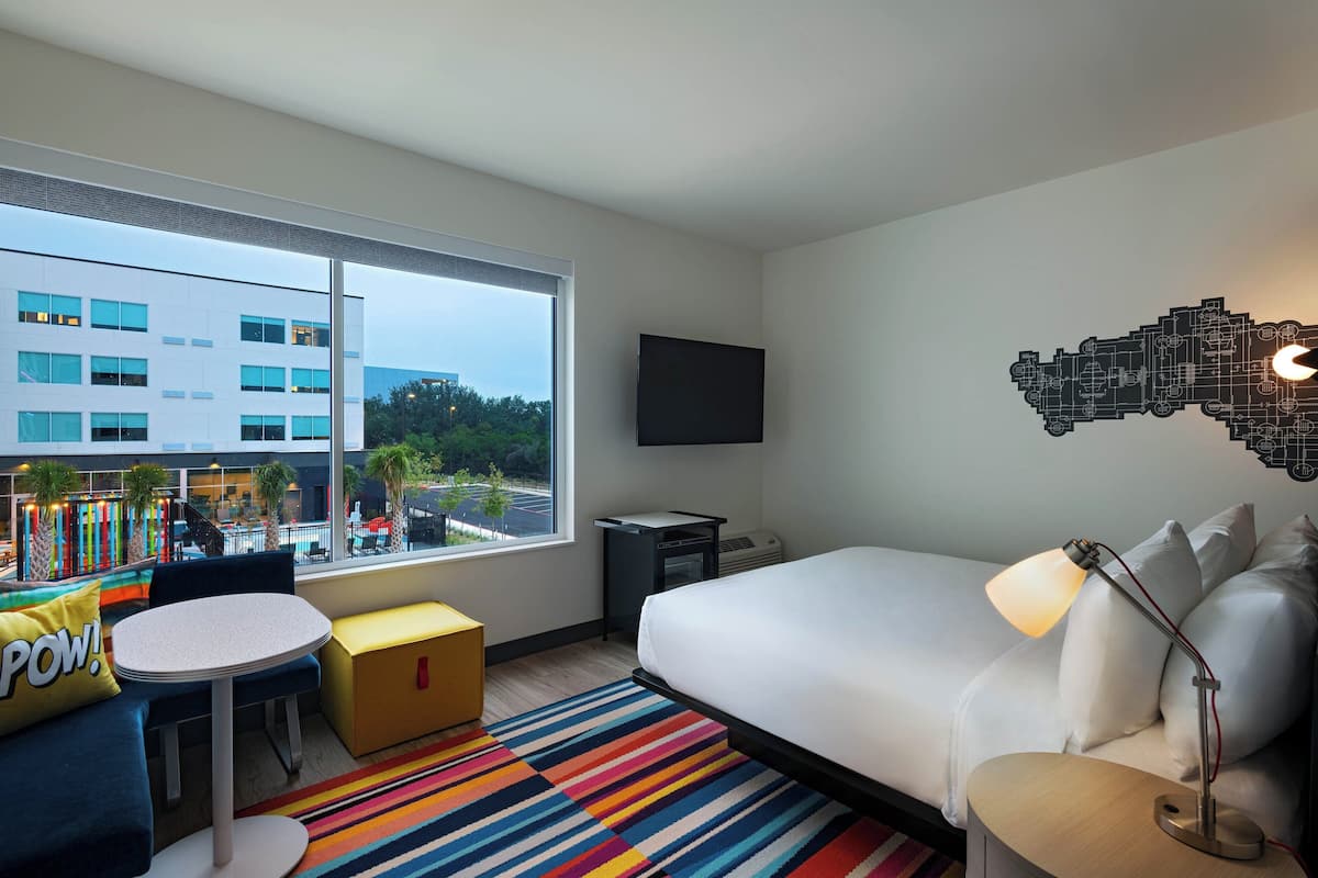 Une chambre d’hôtel avec un grand lit, un bureau, une chaise, une petite table, un tapis rayé aux couleurs vives, un téléviseur à écran plat fixé au mur et une fenêtre donnant sur un immeuble et des arbres.