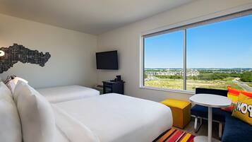 Habitación de hotel con dos camas, televisión, una mesita pequeña, alfombra colorida y un ventanal con vistas a la ciudad.