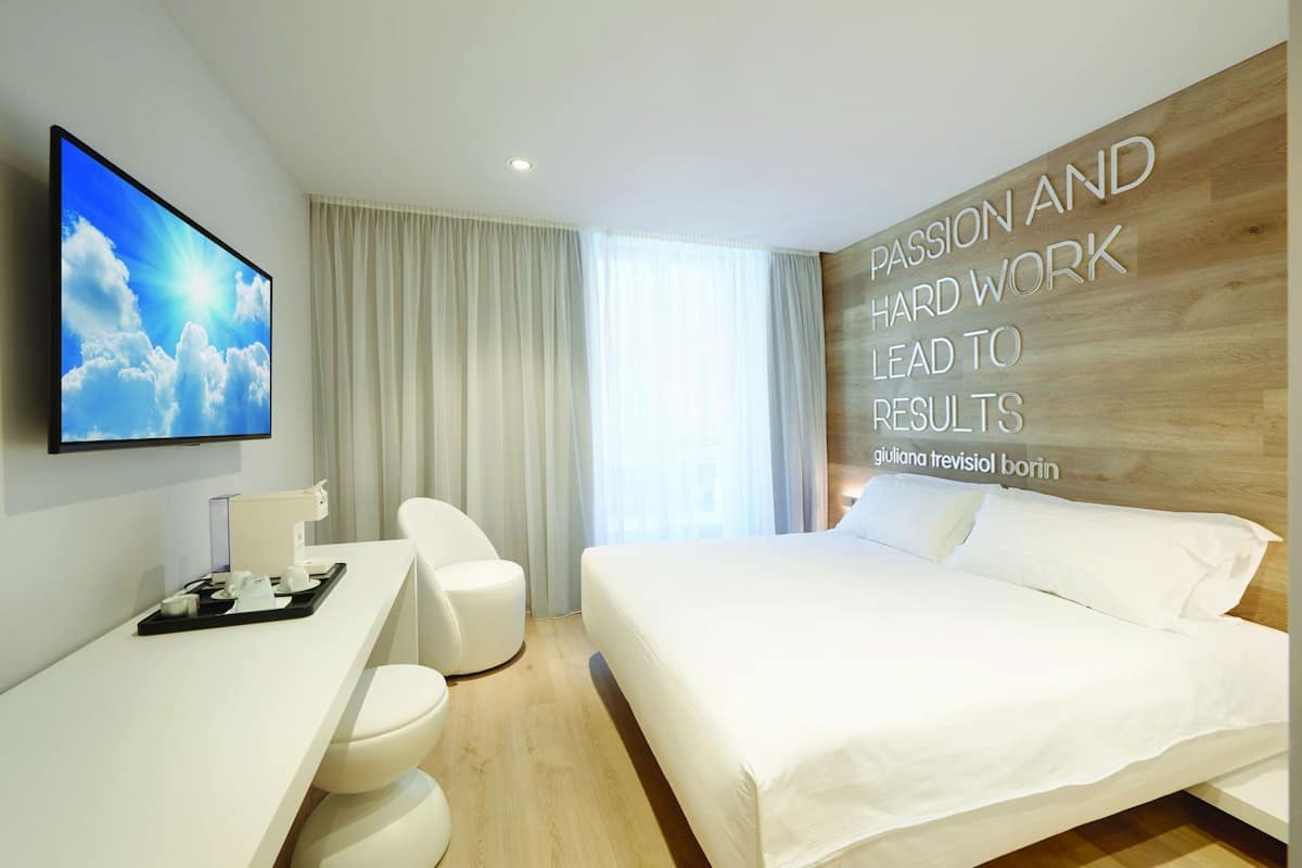 Una habitación de hotel moderna con cama, escritorio con computadora, una silla y un televisor mostrando una escena nublada.