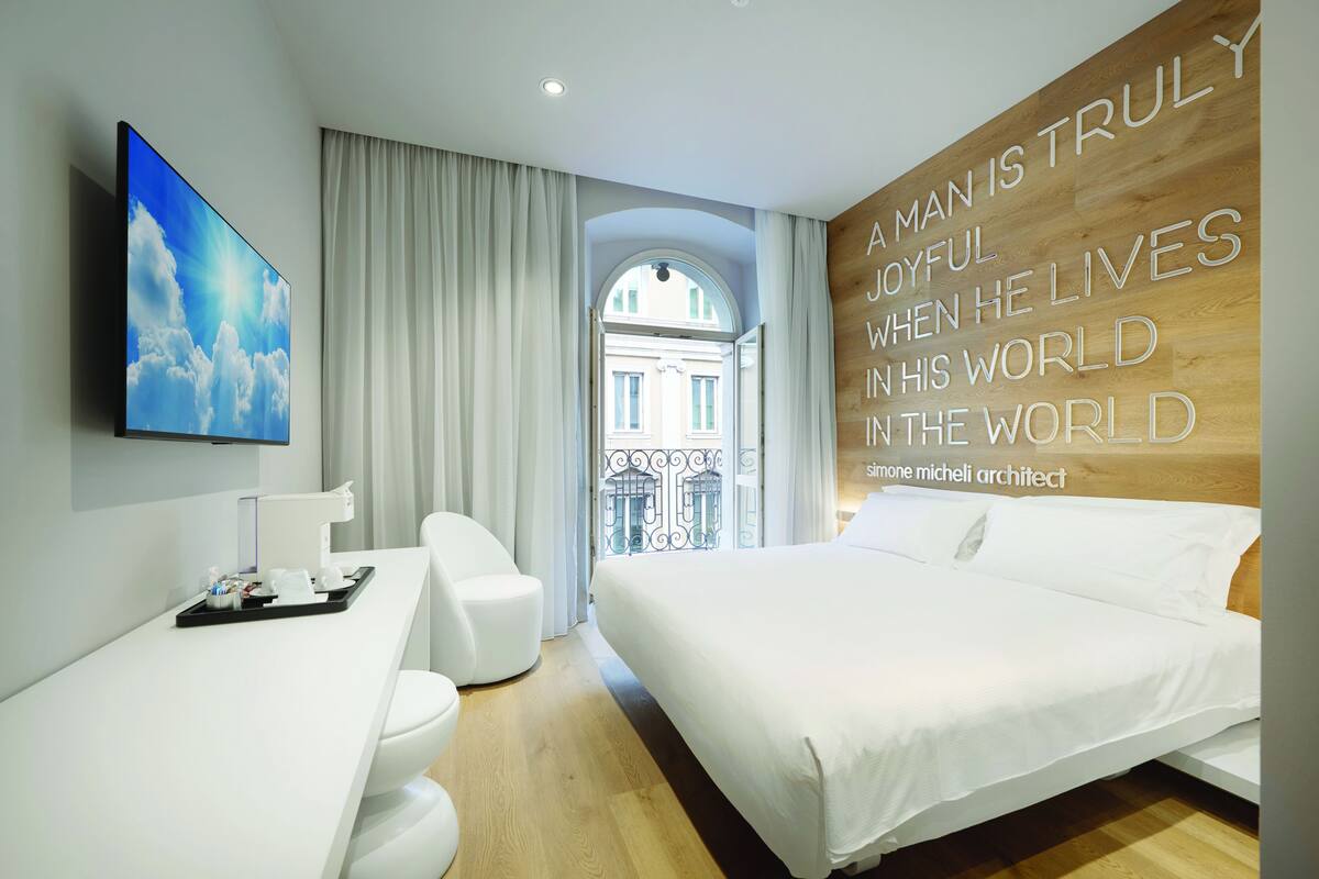 Una habitación de hotel moderna con una cama grande, un escritorio y vistas a un edificio a través de una ventana con arco.