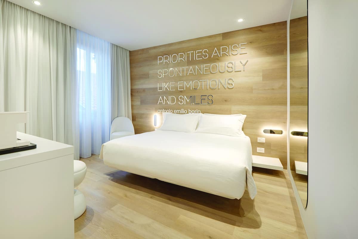 Habitación de hotel moderna con una cama grande, una mesita de noche, un escritorio con silla, un inodoro y un ventanal con cortinas.