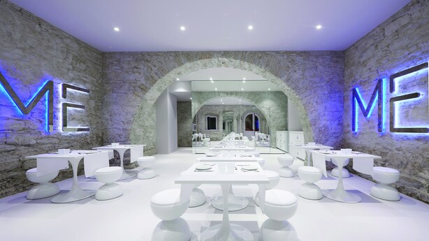 A modern interior with white tables and stools arranged in a spacious room with stone walls and illuminated 'ME' letters. - You.Me Design Place Hotel (Trieste)