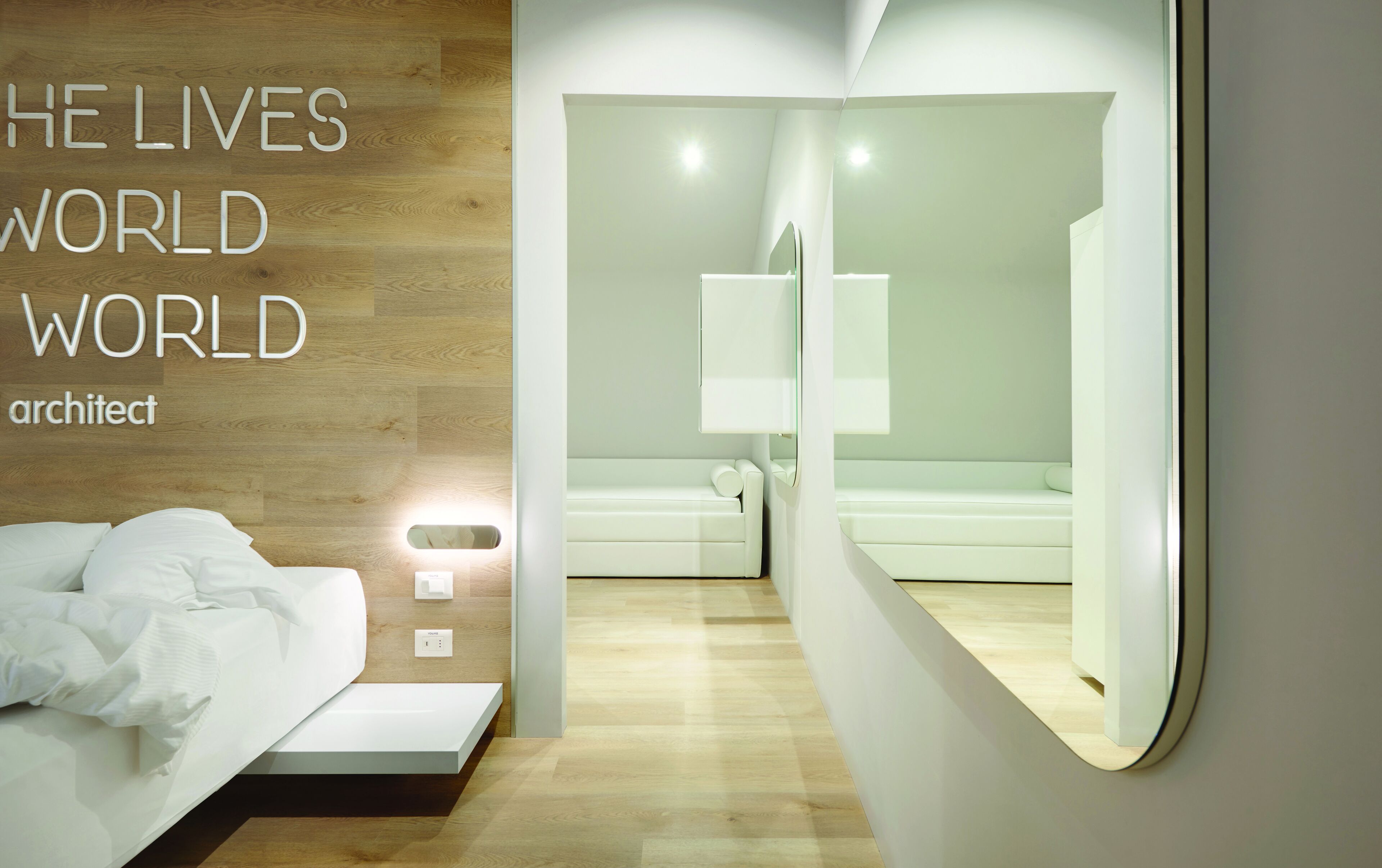 A modern hotel room with a minimalist design, featuring a bed, a large mirror, and a light-colored wooden floor.