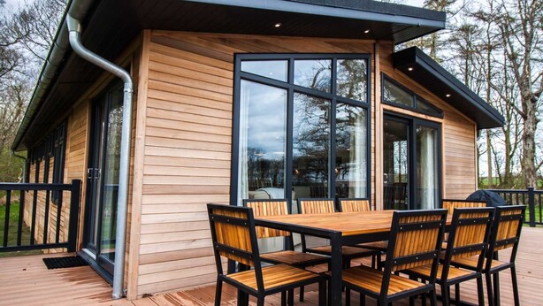 A modern wooden cabin with a deck, large windows, and a dining area with chairs and a table. - Cedar Lodge | sleeps 8 - Hot Tub, Dog Friendly - 5* Gold Award (PICKERING)