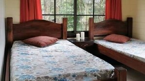 A room with two wooden beds, each with a floral bedspread and a red pillow. There is a window with red curtains, and the room has a tiled floor. - Vistas del Tenorio (Tierras Morenas)