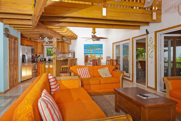 A spacious living area with a large orange sofa, wooden ceiling beams, and a view of the kitchen and outdoor patio.