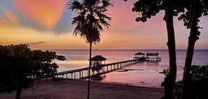 A beach with a wooden pier extending into the water, palm trees, and a sunset.