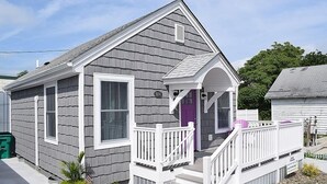 A single-story house with a gray exterior and white trim, featuring a front porch with a white railing and a purple door. - Ivy's Cottage- Renovated, In Town. Great Location. (Chincoteague)