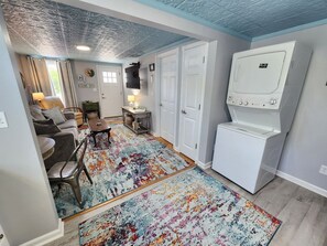 Une buanderie équipée d’un lave-linge et d’un sèche-linge superposés, d’un tapis coloré, d’un canapé, d’une table et d’une porte avec une pancarte.