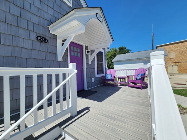 A wooden deck with a white railing, a purple door, and two Adirondack chairs with cushions.